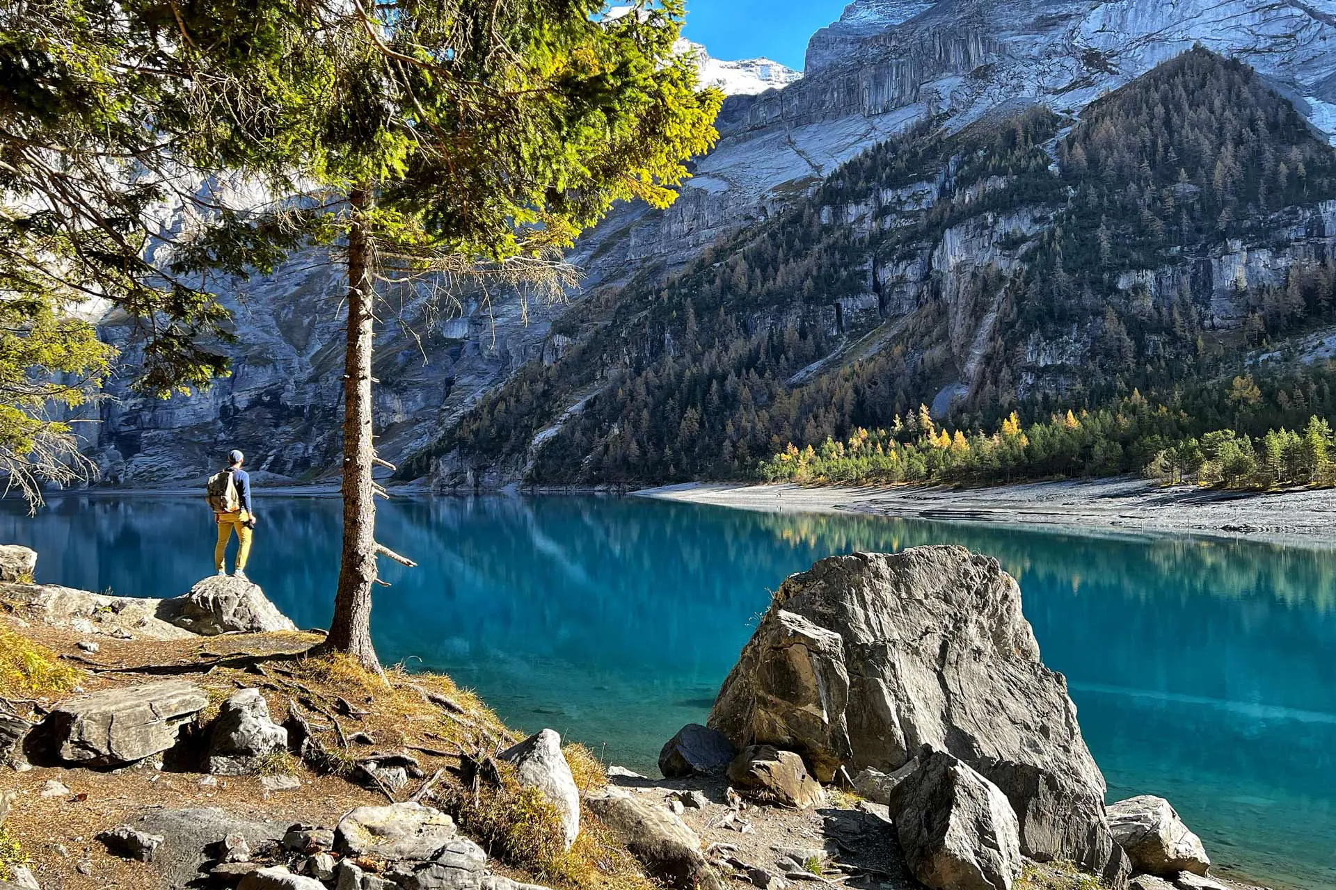 Hiker standing on a rock at the edge of Oeschinensee, looking across the turquoise glacial lake toward the BlĂĽemlisalp limestone cliffs in autumn, Kandersteg Switzerland