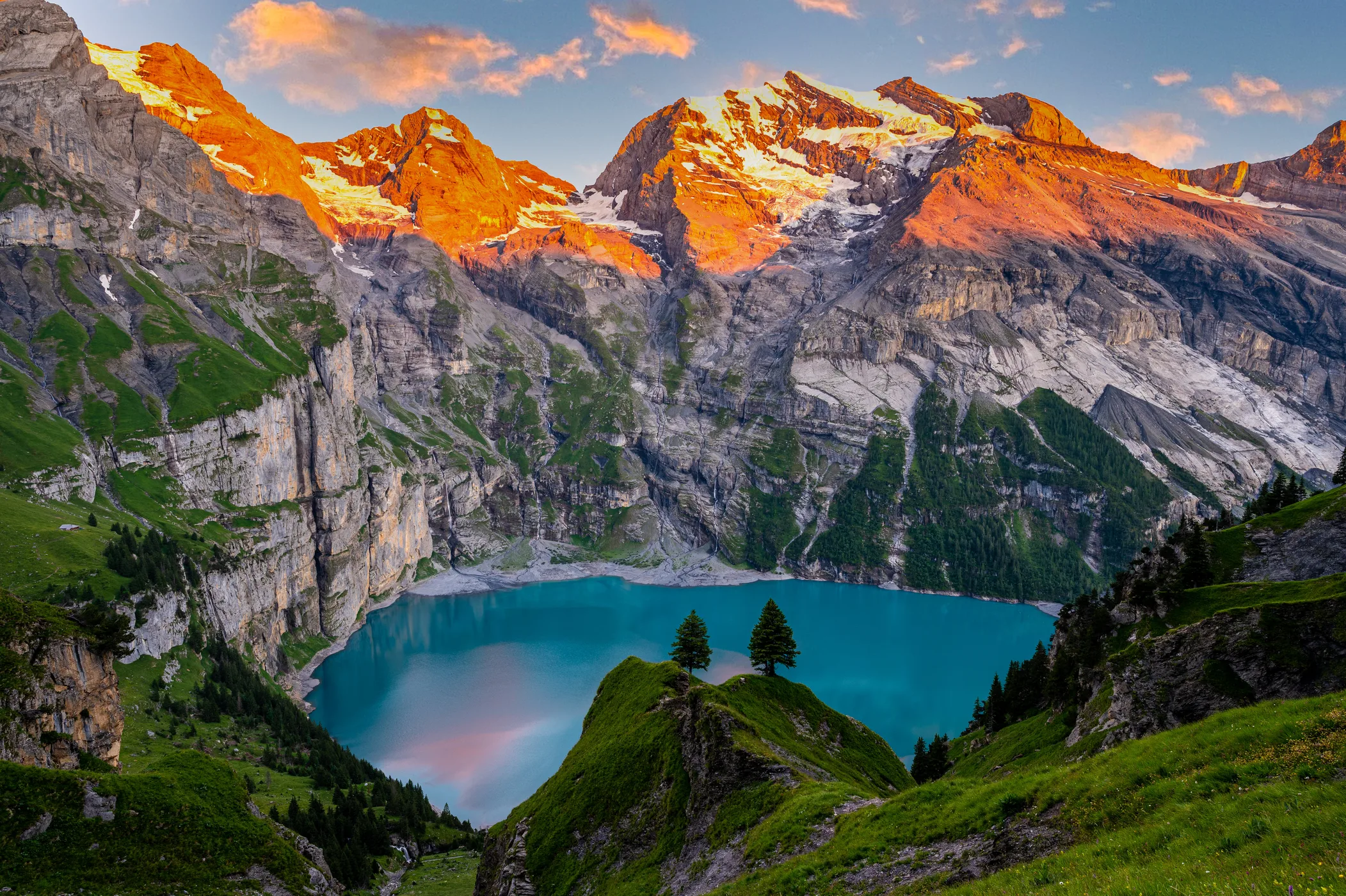 Aerial view of Oeschinensee at sunset with alpenglow turning the BlĂĽemlisalp cliffs orange and gold above the turquoise lake, Bernese Oberland Switzerland