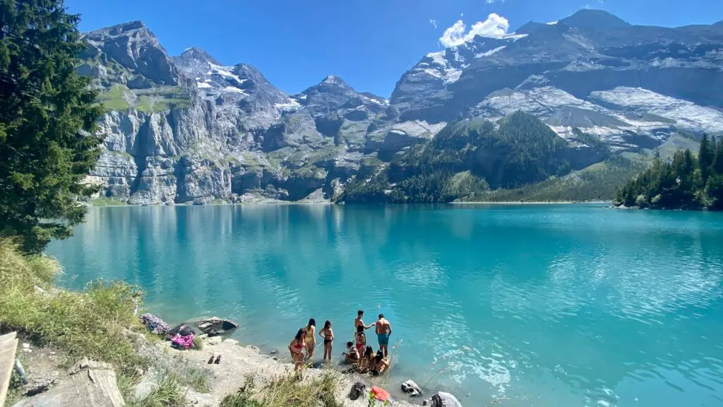 Group of swimmers at the shore of Oeschinensee on a summer day, with the full turquoise lake and BlĂĽemlisalp mountain peaks in the background, Kandersteg Switzerland
