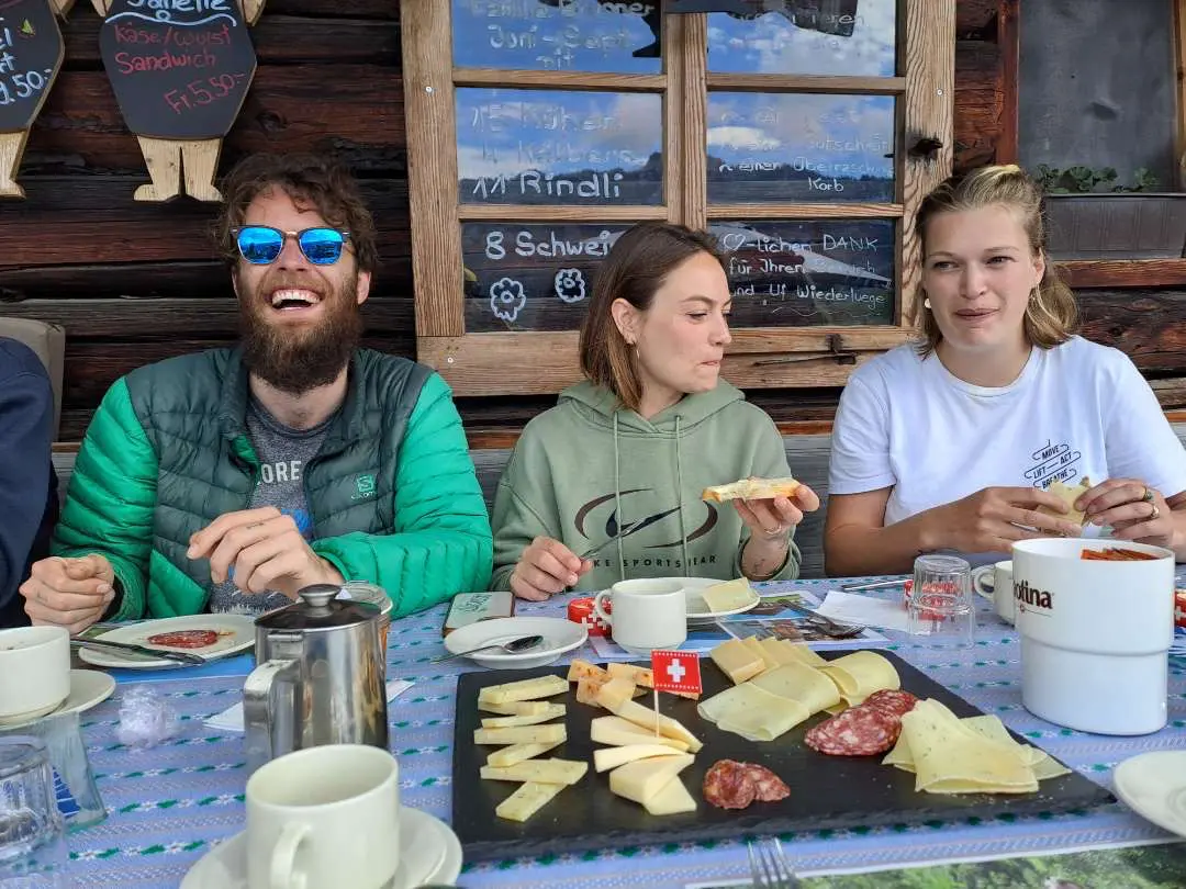 Guests laughing over an alpine farm breakfast with fresh cheese platter and charcuterie outside at Alp Signswiler, Justistal, farm tour from Interlaken