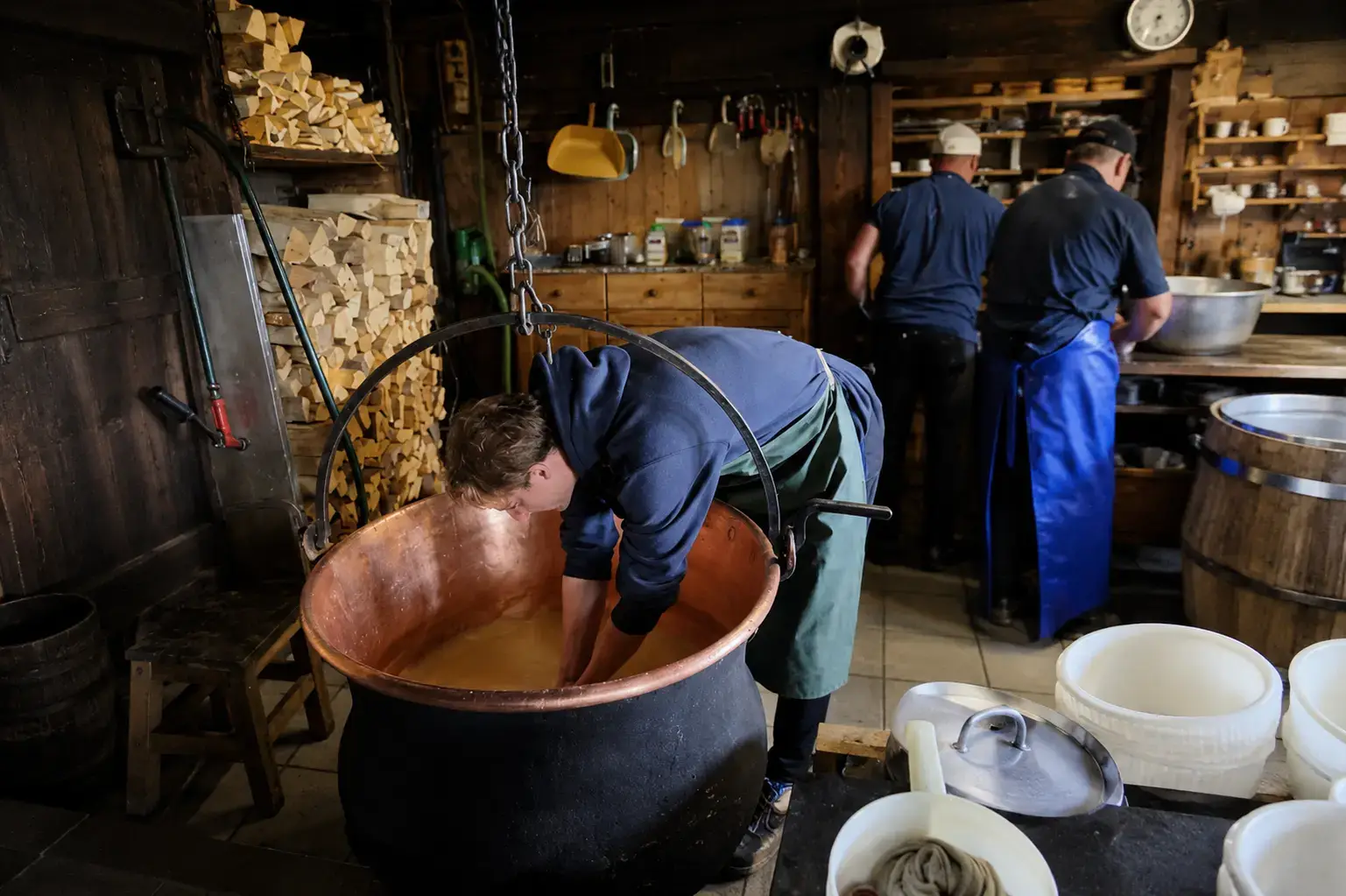 Traditional Swiss cheese makers stirring milk in a copper cauldron over fire inside a rustic alpine farm dairy
