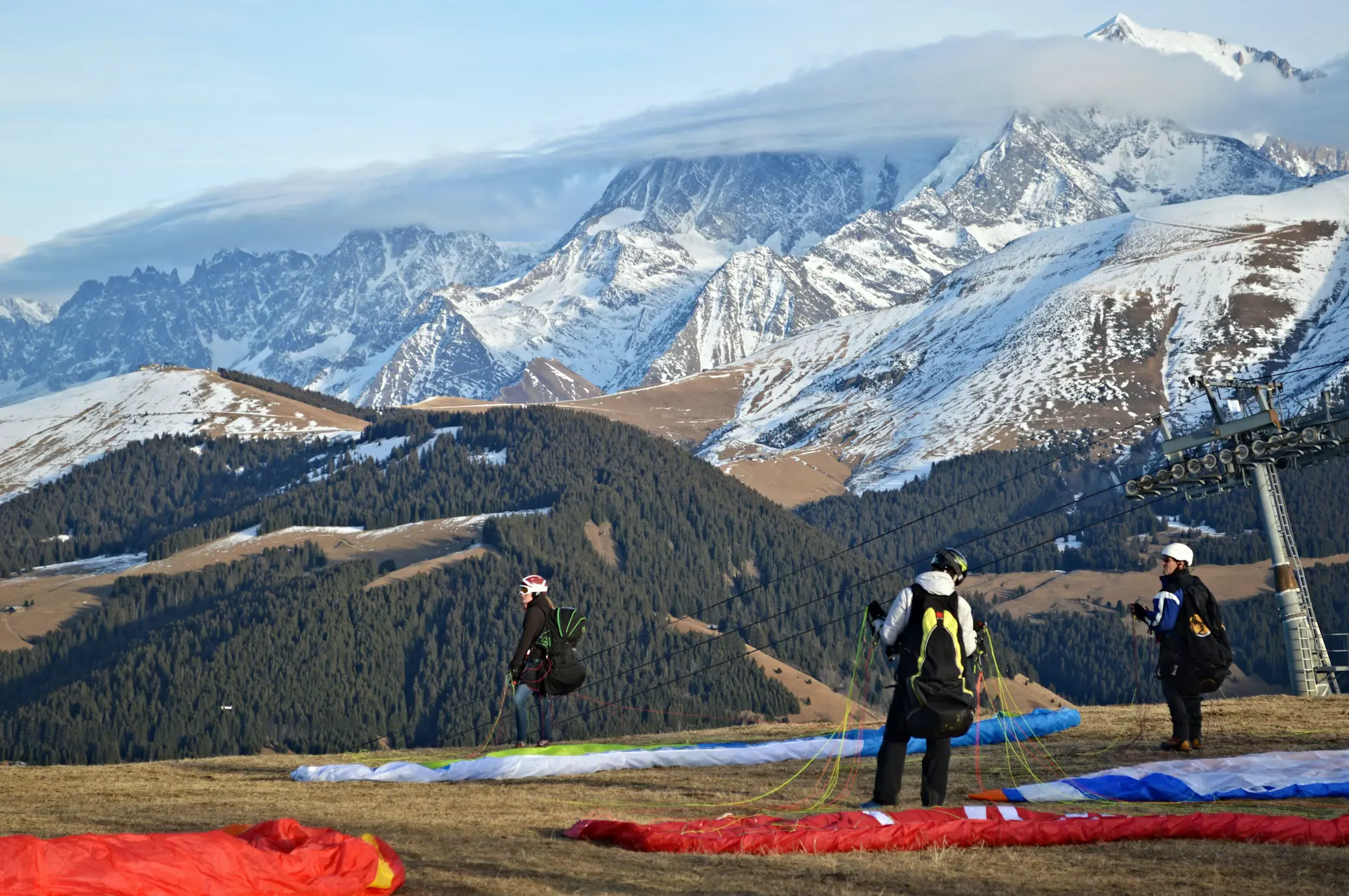 Paragliding pilots preparing canopies on alpine launch site with snow-capped Swiss Alps peaks and cable car in background