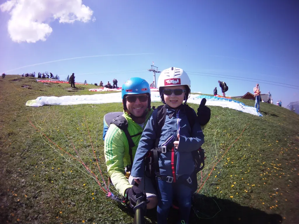Young child with thumbs up in paragliding harness with pilot at alpine launch site, children tandem paragliding Switzerland