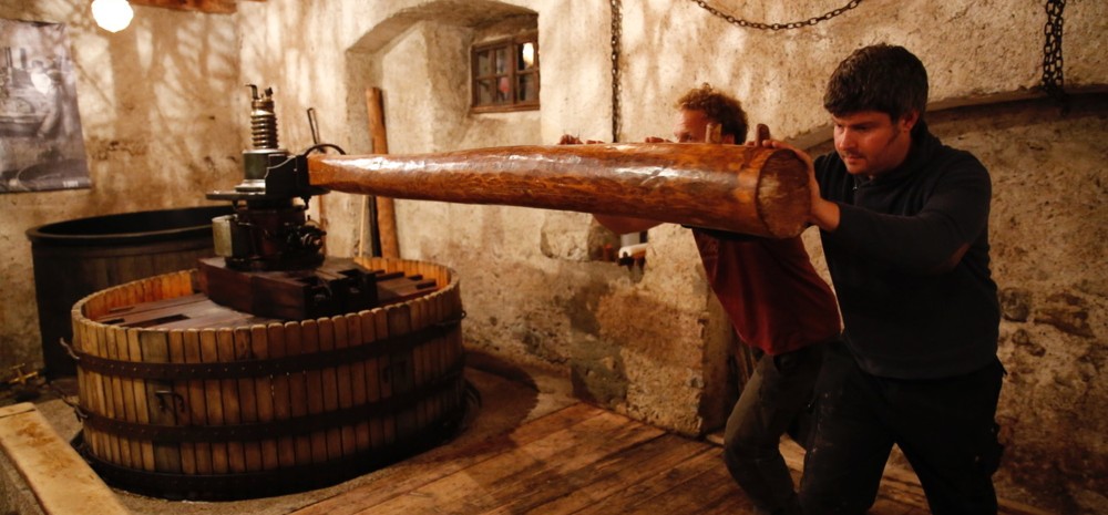 Two winemakers operating a traditional wooden grape press in a stone cellar, ancestral Swiss winemaking technique