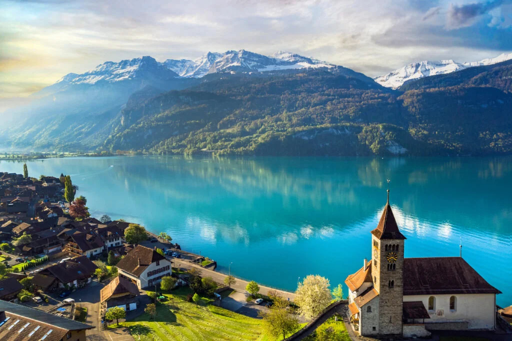 Alpine village with stone church on turquoise Lake Brienz shore and snow-capped mountains near Interlaken Switzerland