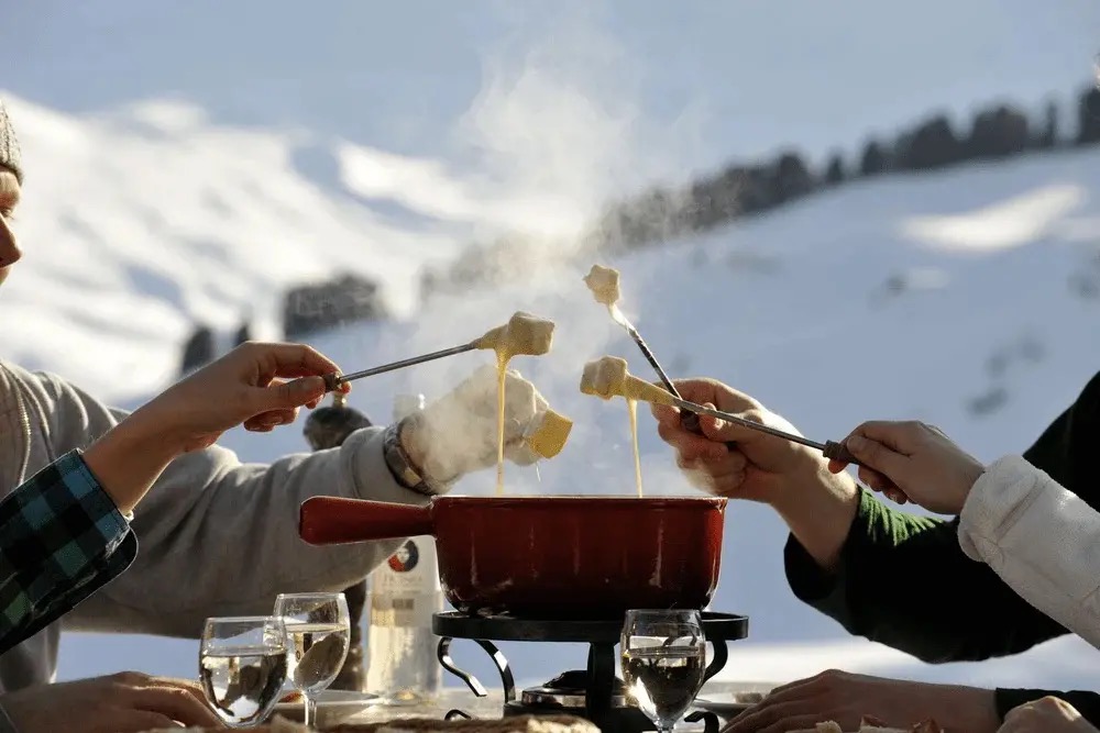Group sharing Swiss cheese fondue outdoors with bread on long forks steaming red pot and snowy alpine slopes Interlaken