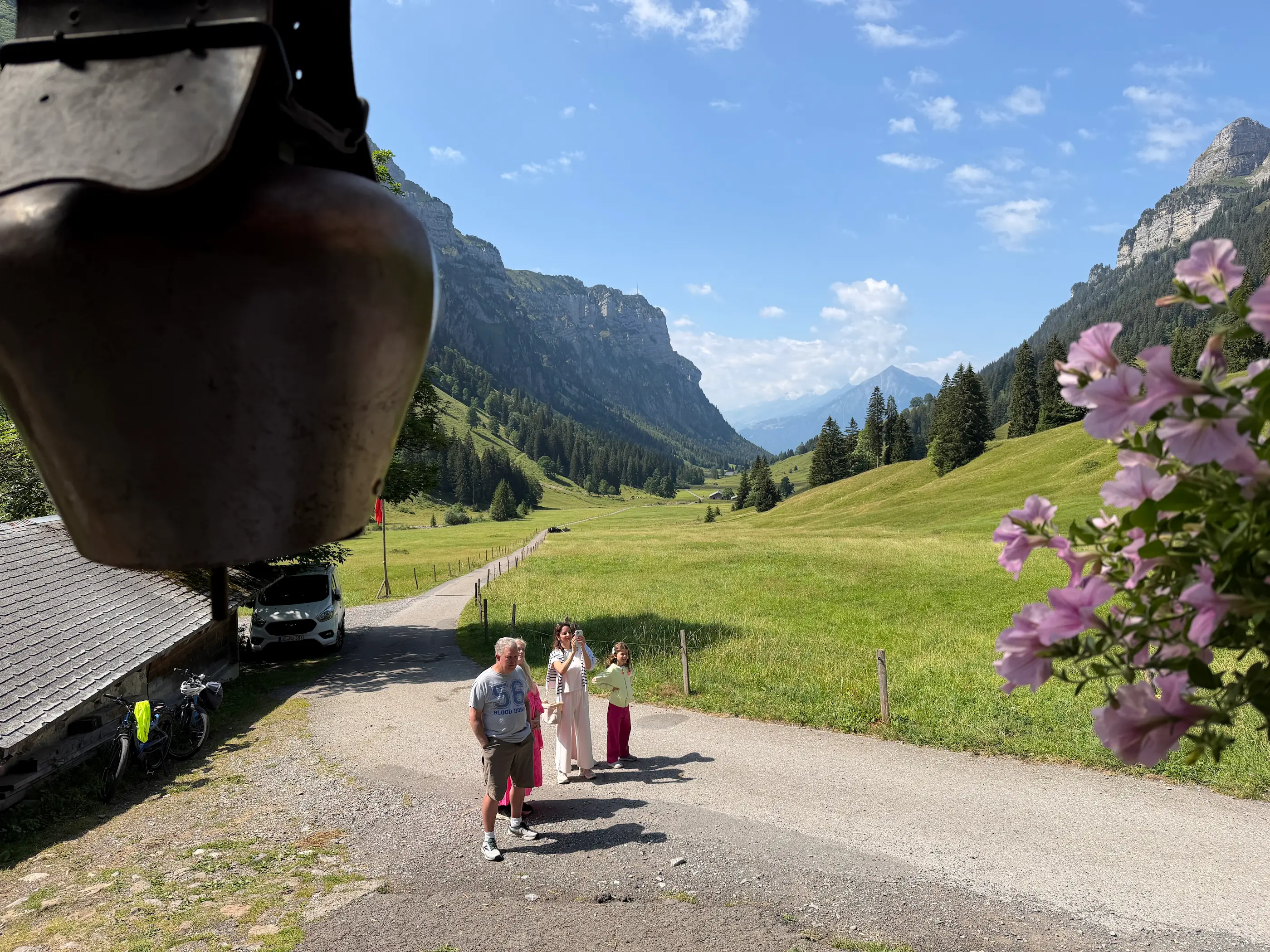 Family arriving at the alpine farm in the Justistal, framed by a hanging cowbell and pink flowers, with the valley and limestone cliffs behind them