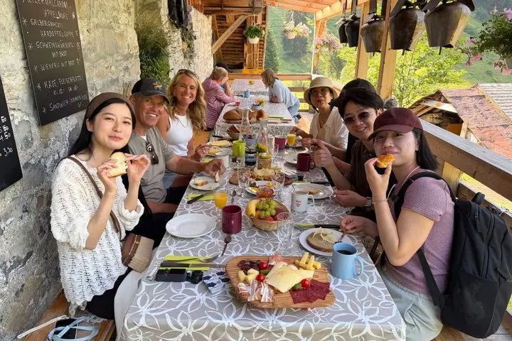 Farm Tour guests sharing a Swiss farm breakfast on the covered terrace, with boards of alpine cheese and cured meat, fresh bread and cowbells hanging overhead