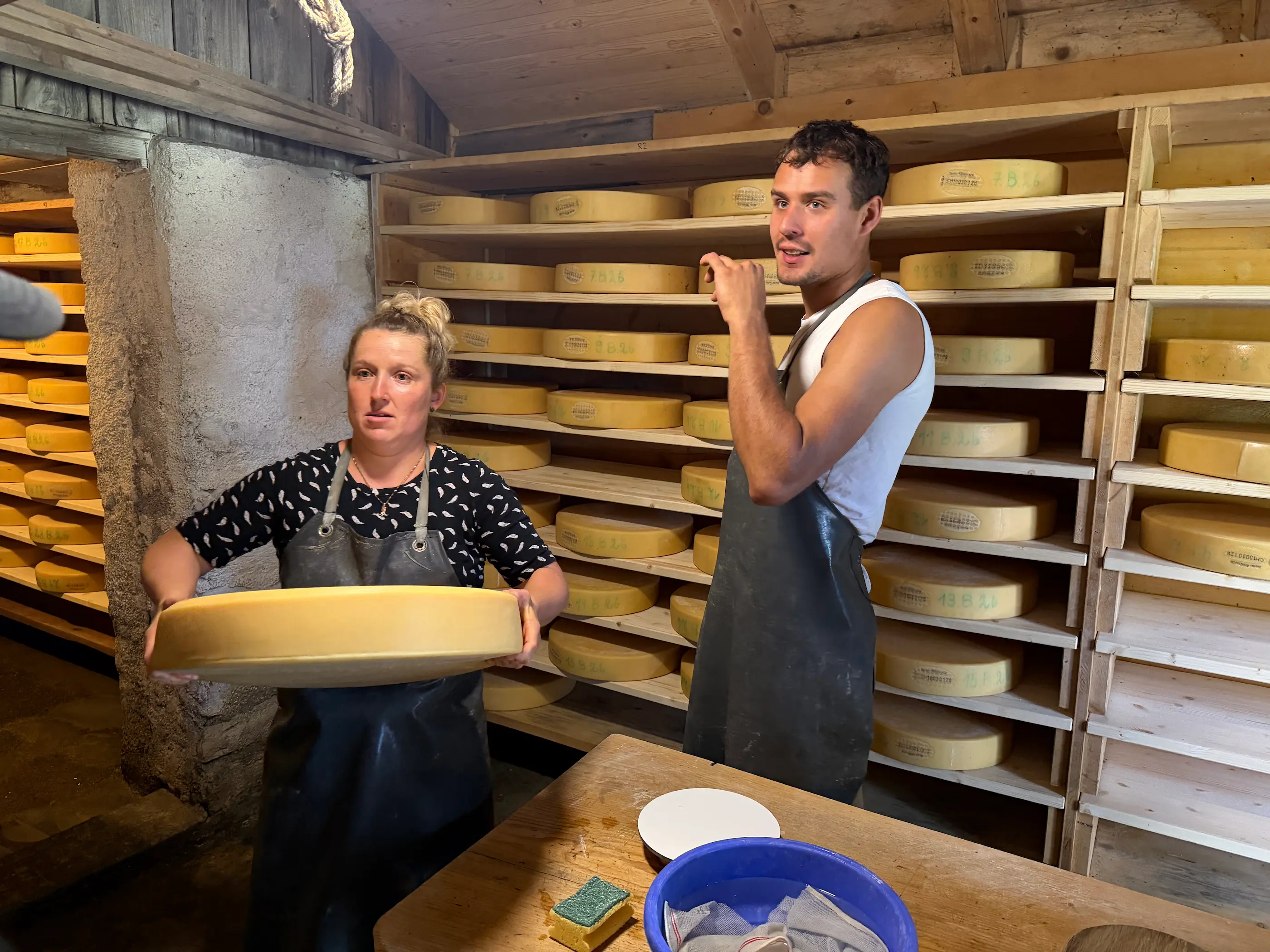 Two alpine cheesemakers in the farm's ageing cellar, one holding a wheel of alpine cheese in front of wooden shelves stacked with maturing wheels