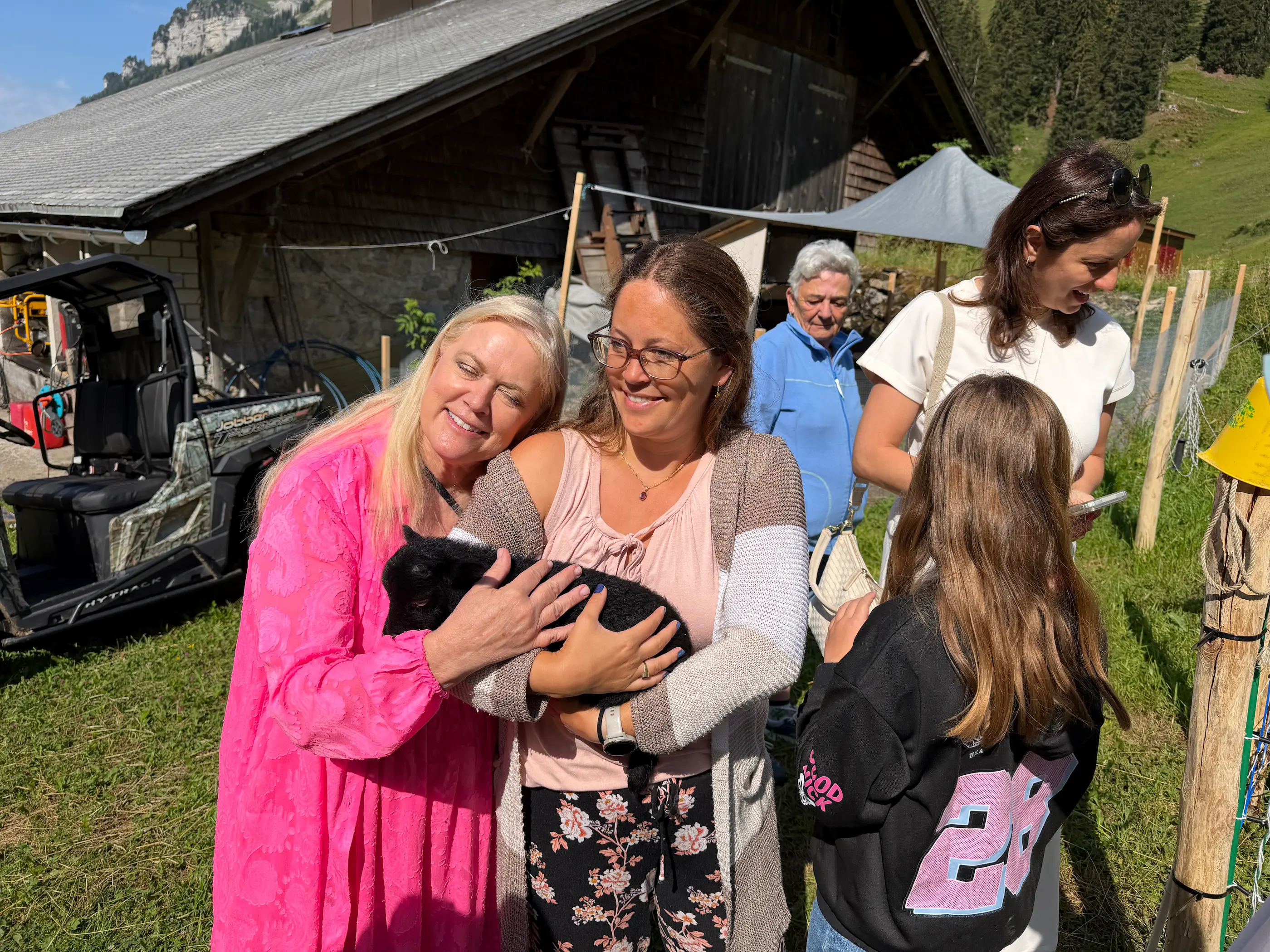 Farm Tour guests smiling as they hold a black lamb outside the farmhouse, with the rest of the small group watching in the morning sun