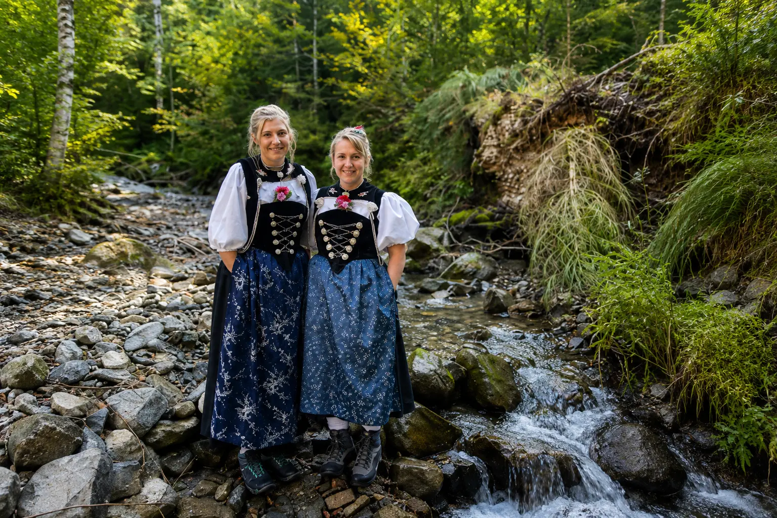 Two Swiss women in traditional Bernese Tracht dresses standing on the rocks beside a mountain stream in the forest