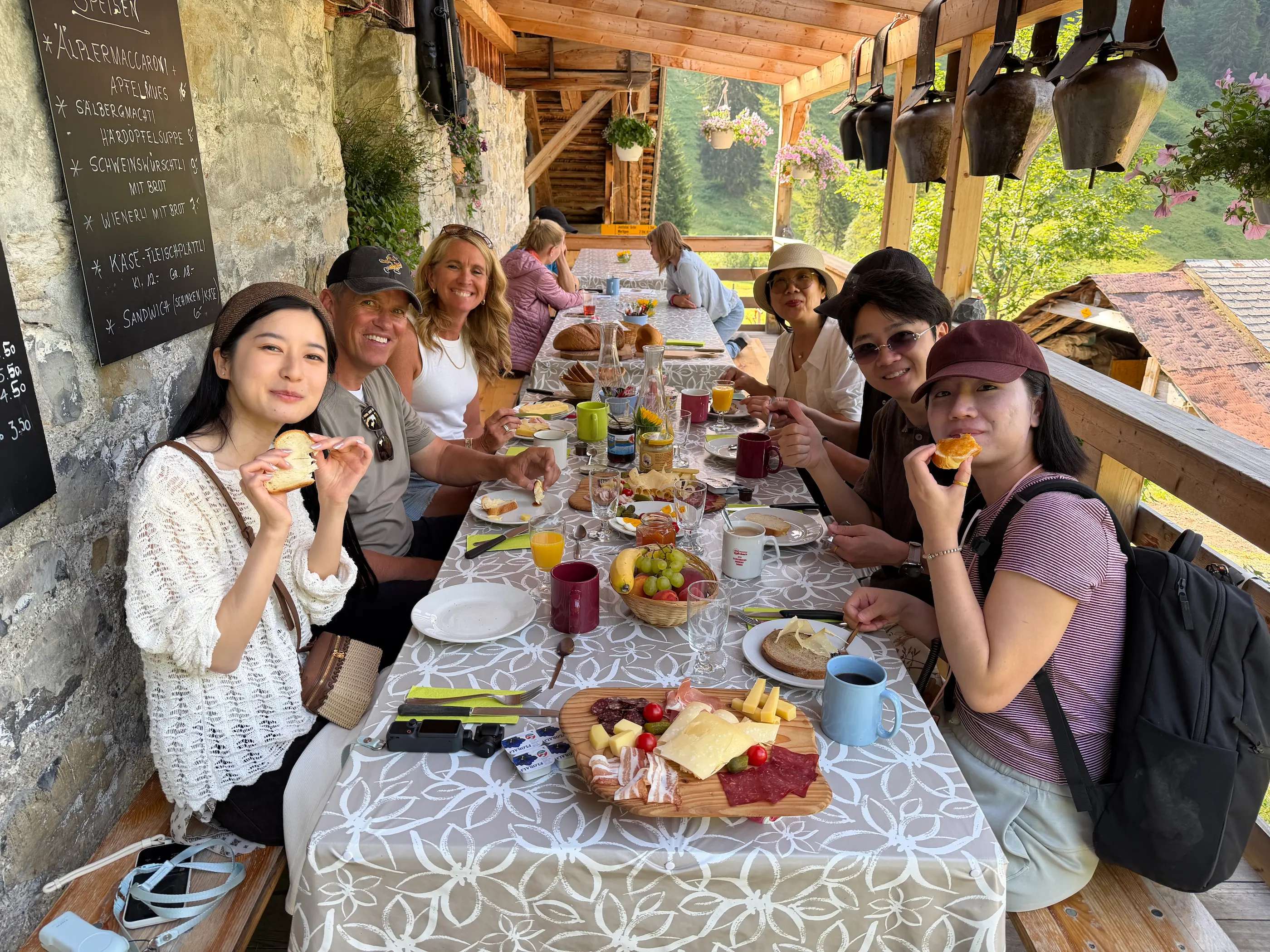 Guests sharing an alpine farm breakfast on a Swiss farm terrace, cheese, cured meat and bread from the same valley
